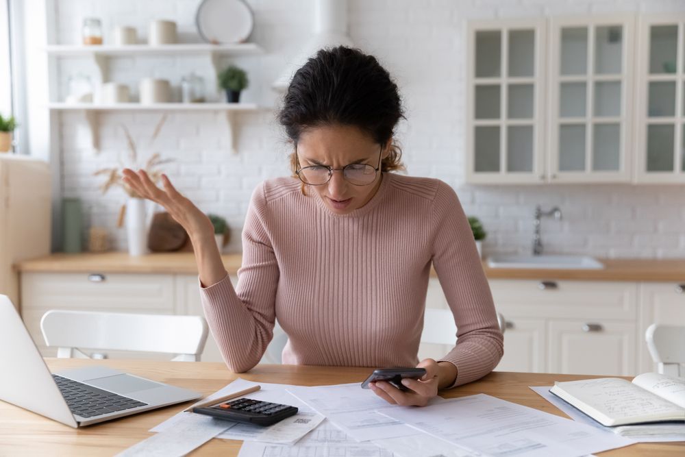 Person working at a kitchen table, looking frustrated while using a phone beside papers and a laptop.