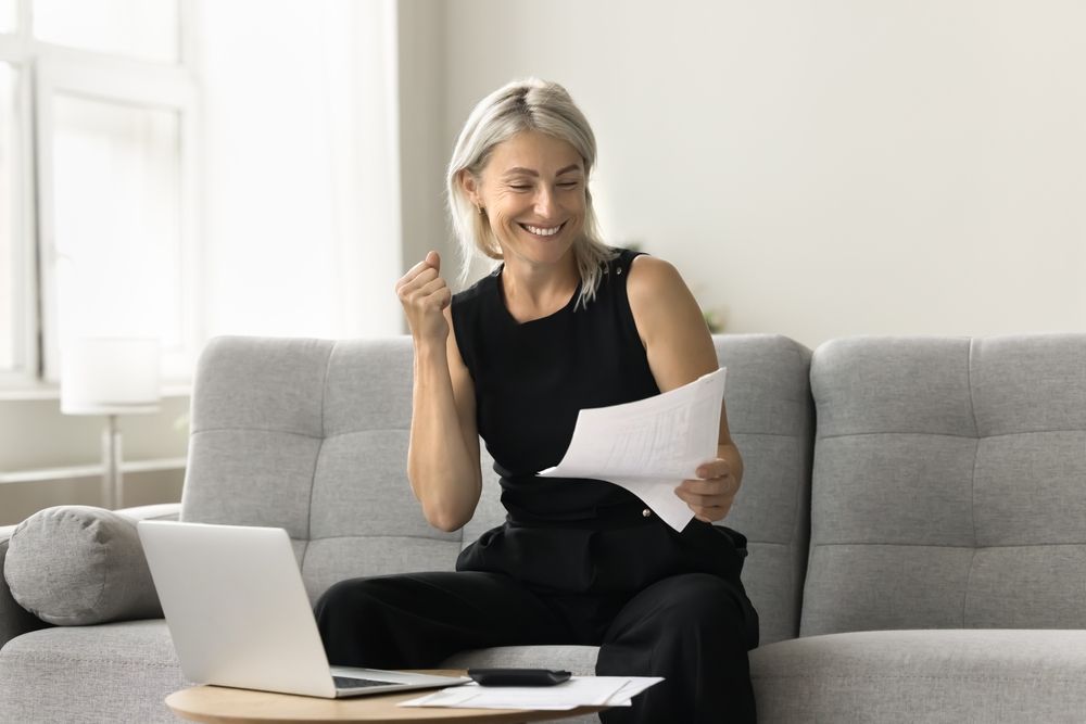 Woman on a gray sofa smiling at a laptop while holding a sheet of paper