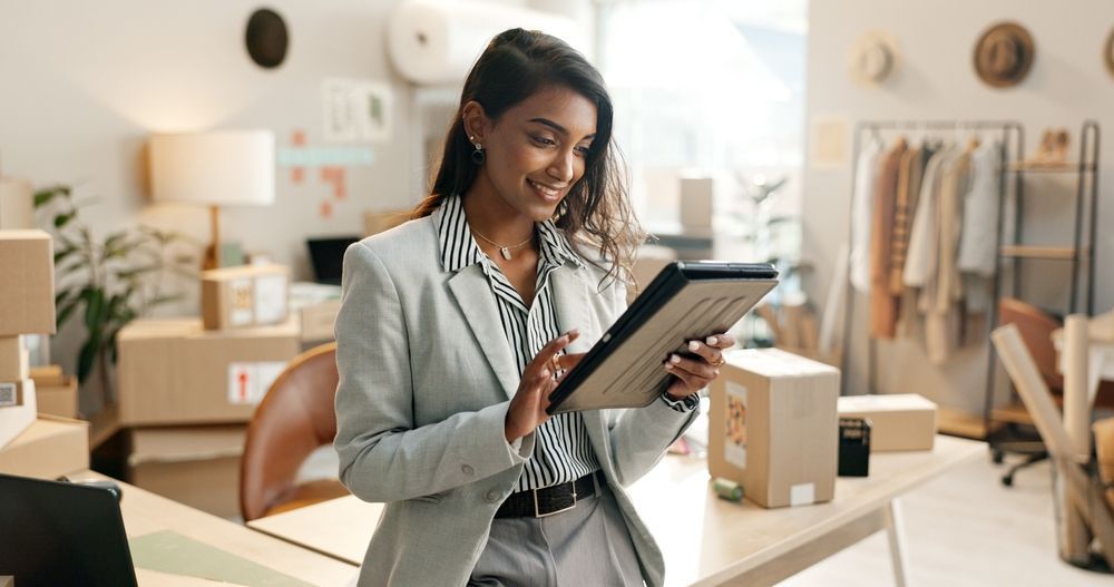 Woman in a light gray blazer using a tablet in a bright boutique workspace with boxes and clothing racks