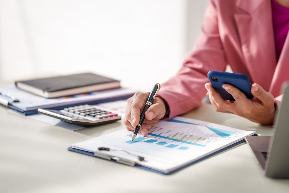 Person in a pink shirt reviewing charts and writing on a clipboard while holding a phone at a desk