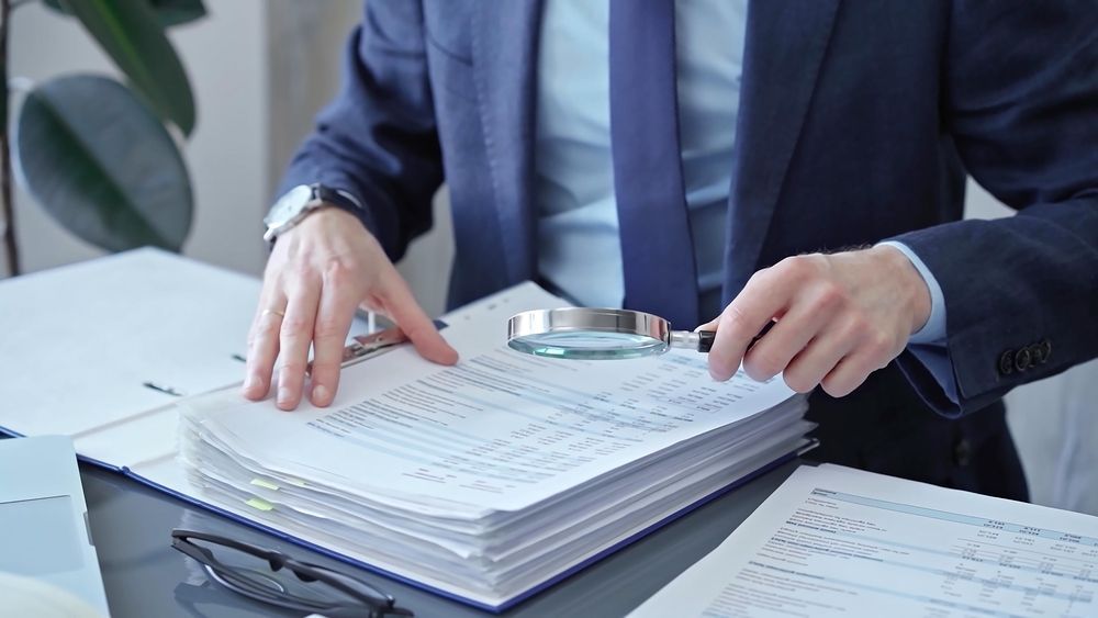 Person in a dark suit stamping documents on a desk with stacked papers and a binder in an office.