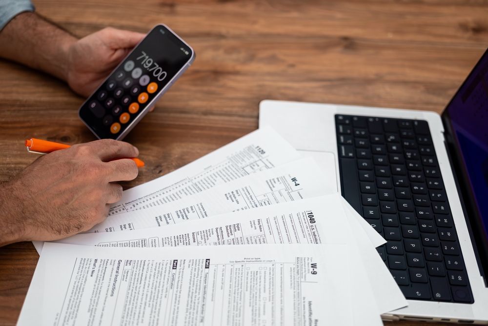 Hands using a calculator over paperwork beside a laptop on a wooden desk