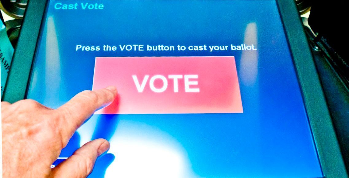 Finger pressing pink “VOTE” button on a blue touchscreen voting machine with ballot instructions.