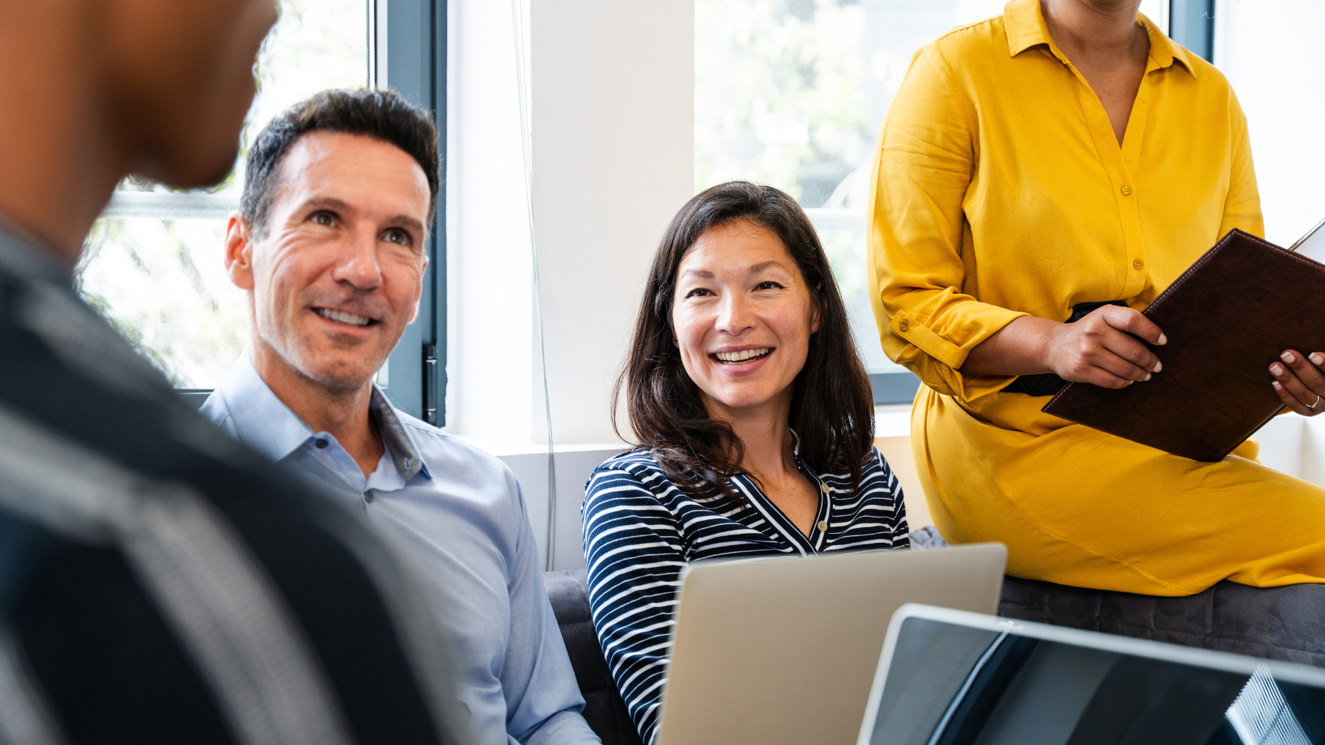 Three coworkers in an office meeting, smiling with a laptop and clipboard.