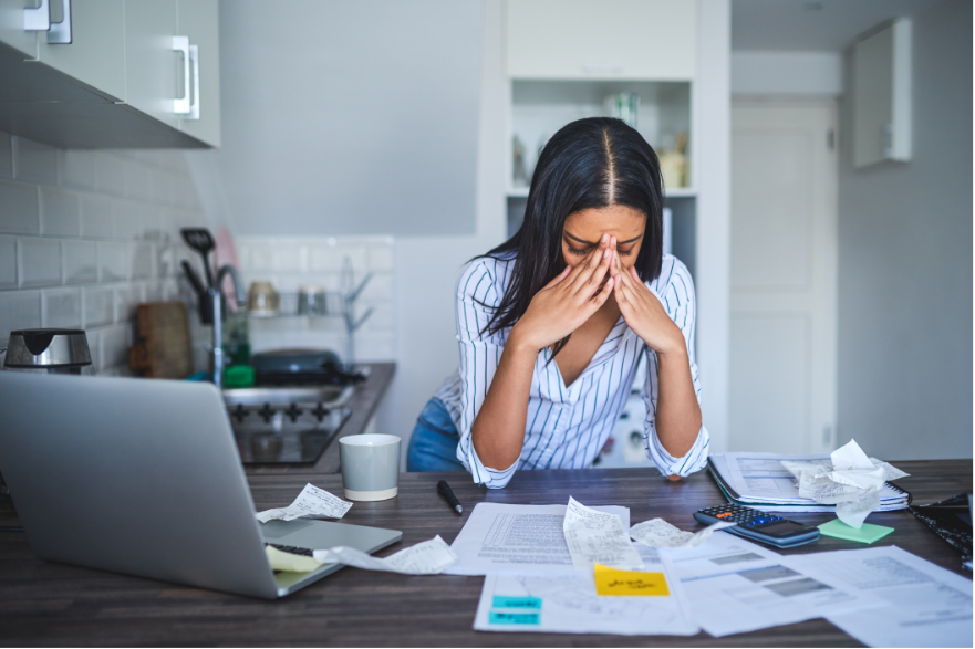 Person sitting at a cluttered desk, stressed with head in hands, surrounded by papers and a laptop in a home office