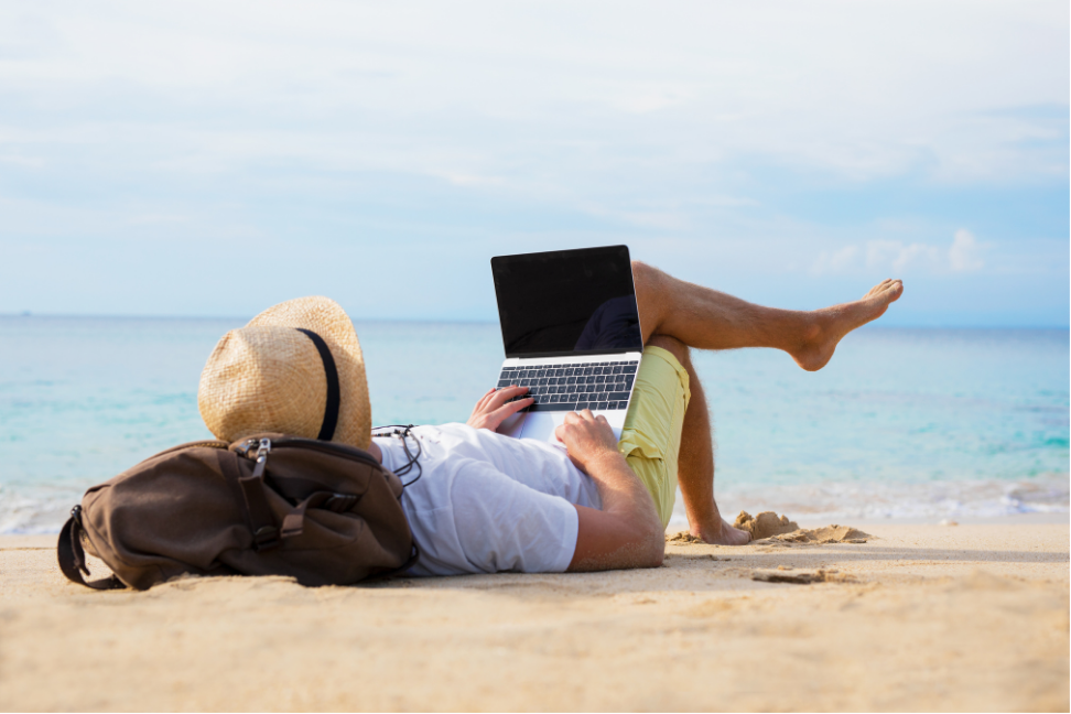 Person lounging on a sandy beach with a laptop, backpack, and sunhat by the ocean