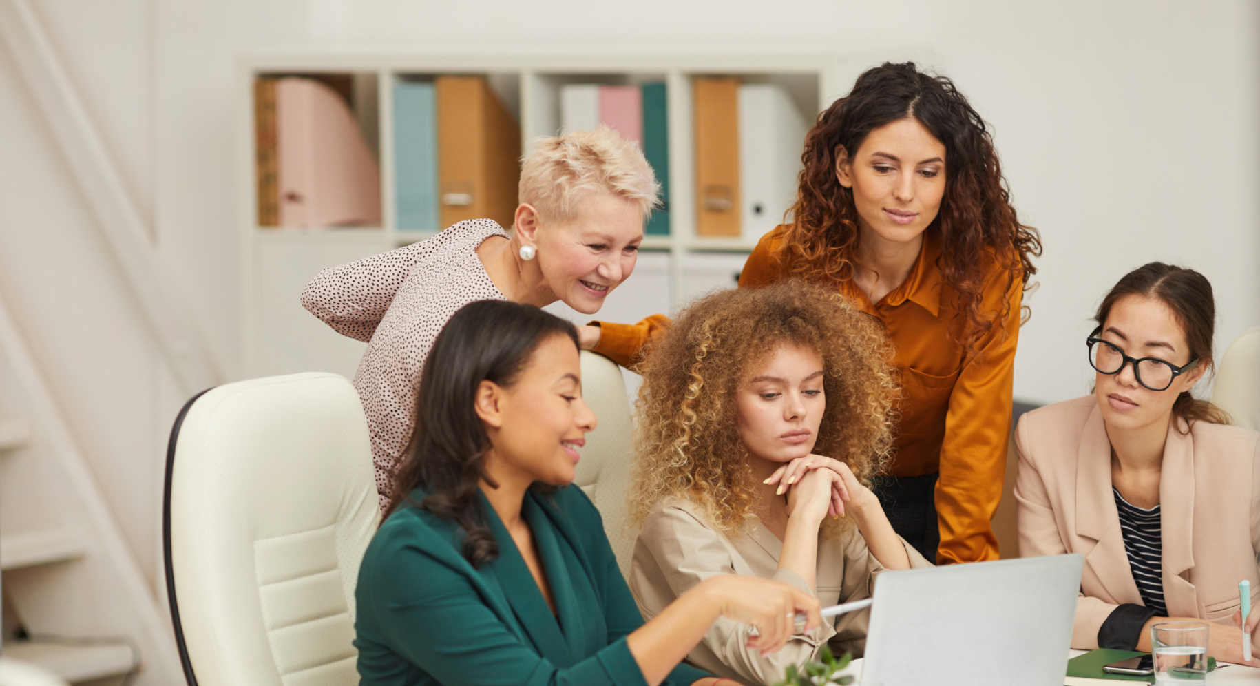 Five coworkers gathered around a laptop in a bright office, discussing a project together.