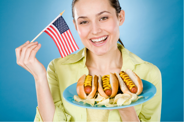 Smiling woman holding a plate of hot dogs and a small American flag on a blue background