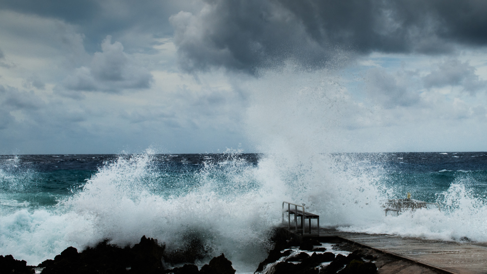 Large waves crash over a rocky seawall and pier under a cloudy sky.