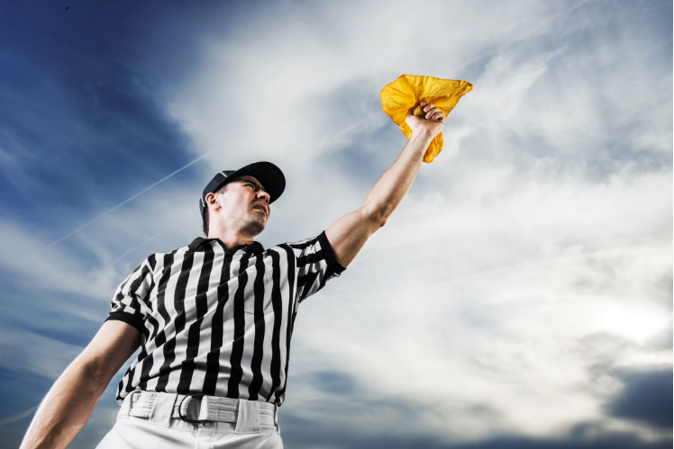 Referee in black-and-white stripes raises yellow penalty flag against a cloudy sky
