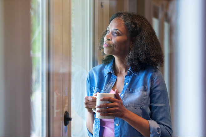 Woman in a blue shirt holding a mug by a window, gazing outside with a calm expression