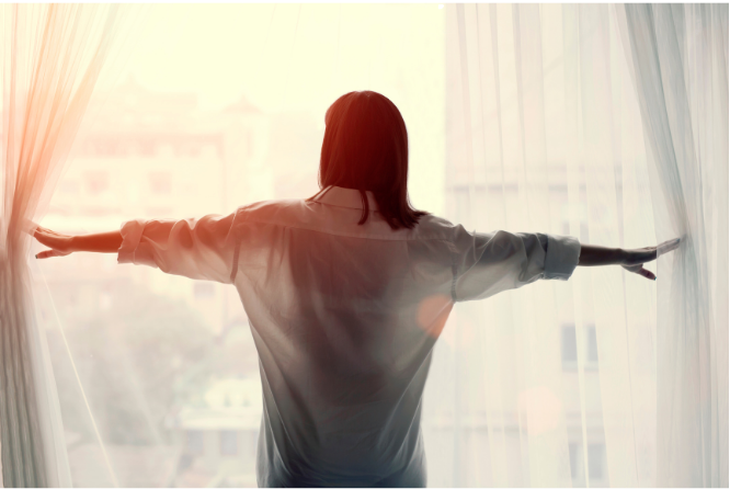 Person standing with arms outstretched before bright curtains, backlit by sunlight