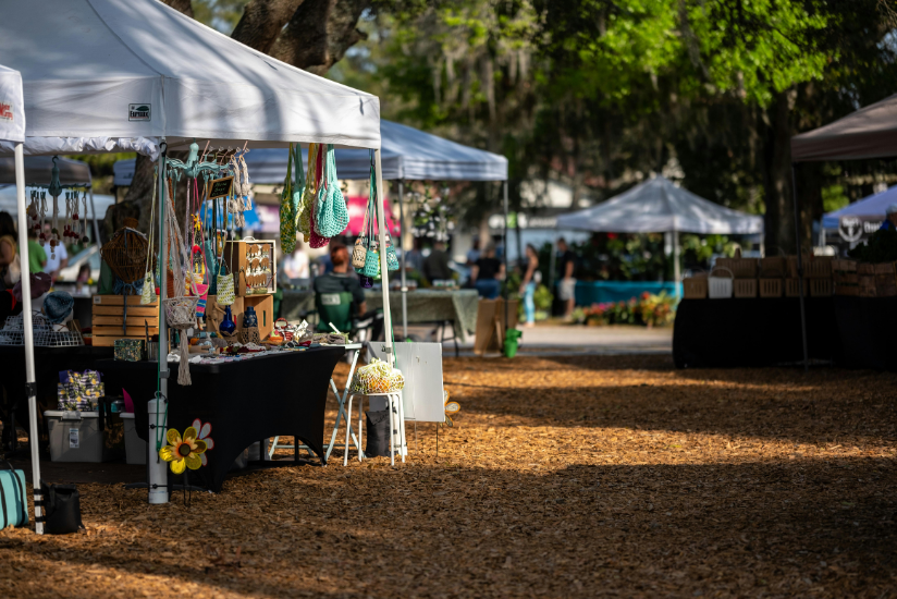 Outdoor market stalls under white tents in a shaded park, with handmade goods displayed on tables.