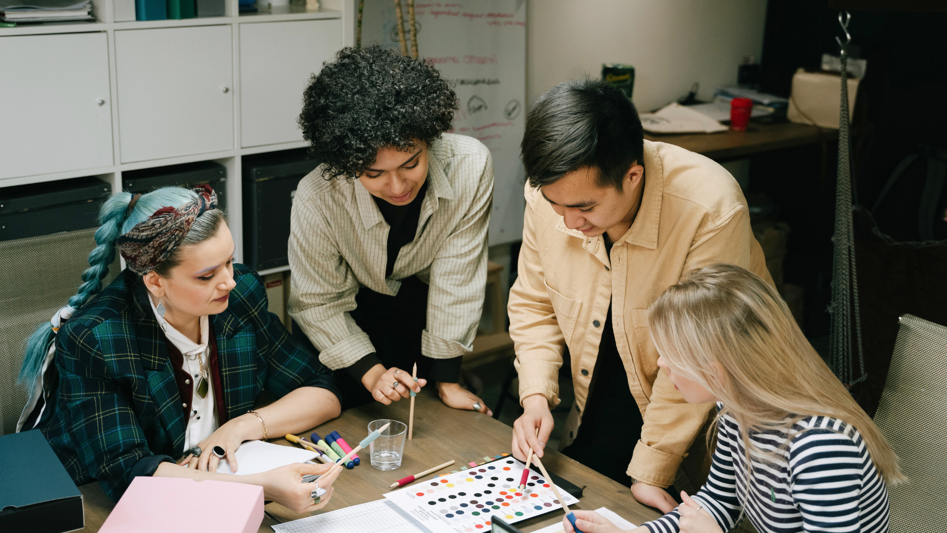 Four coworkers lean over a table, discussing colorful papers and markers in an office.