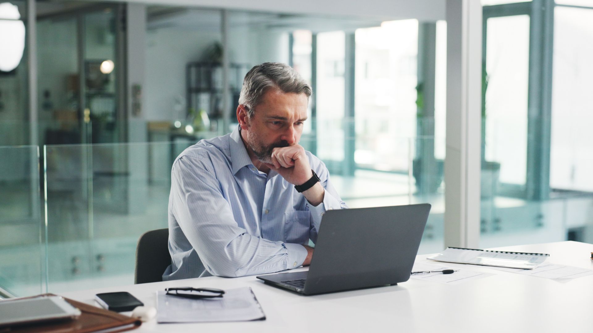 Man in a glass office reviewing a laptop, hand on chin, with papers on the desk