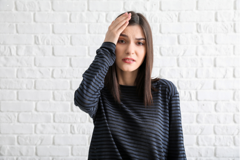 Woman with hand on forehead, standing against a white brick wall, looking concerned
