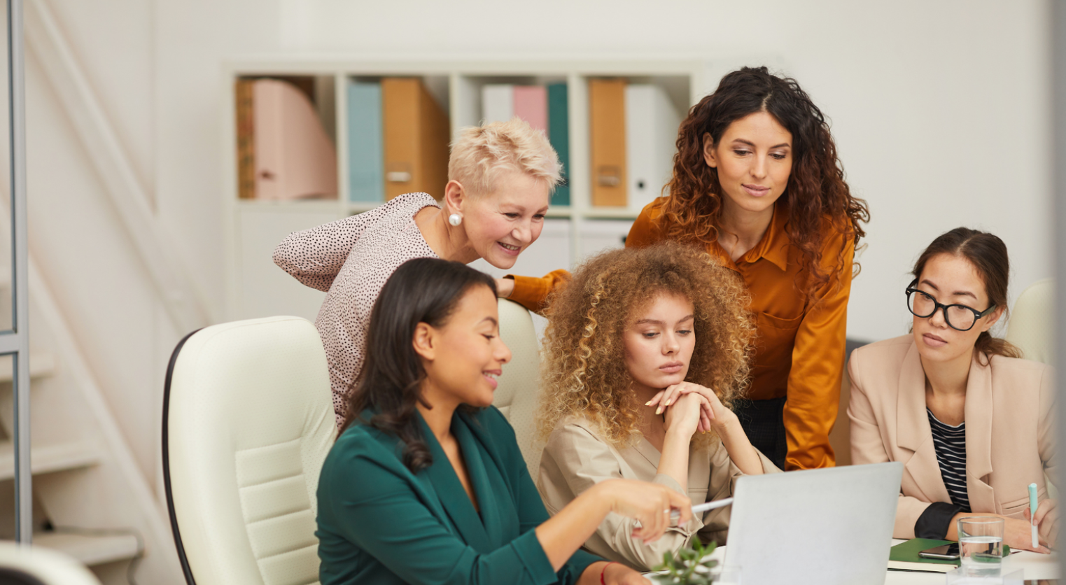 Five coworkers gathered around a laptop in a bright office, collaborating and looking at the screen.