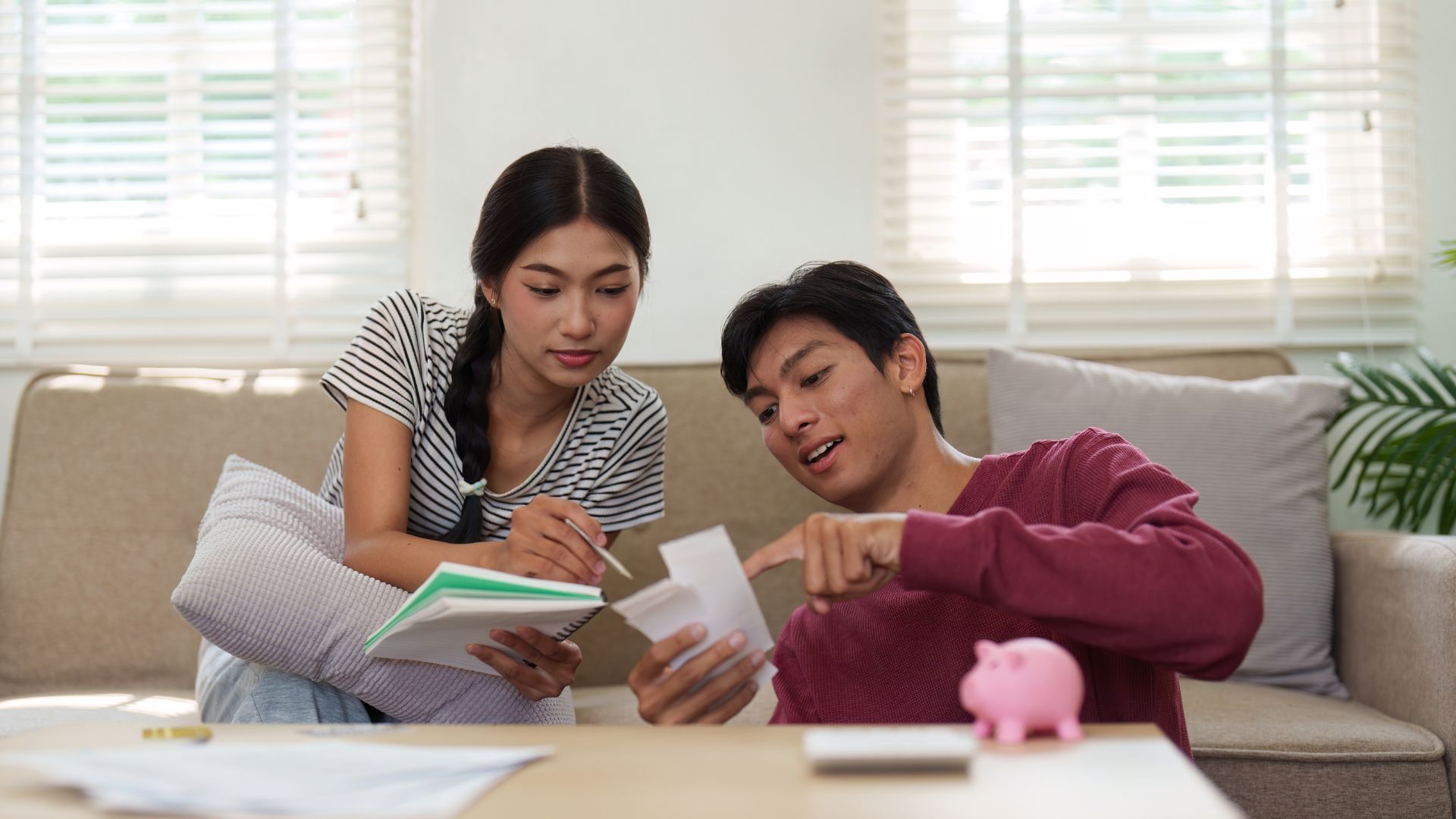 Two people on a couch reviewing papers in a bright living room, with a piggy bank on the table.