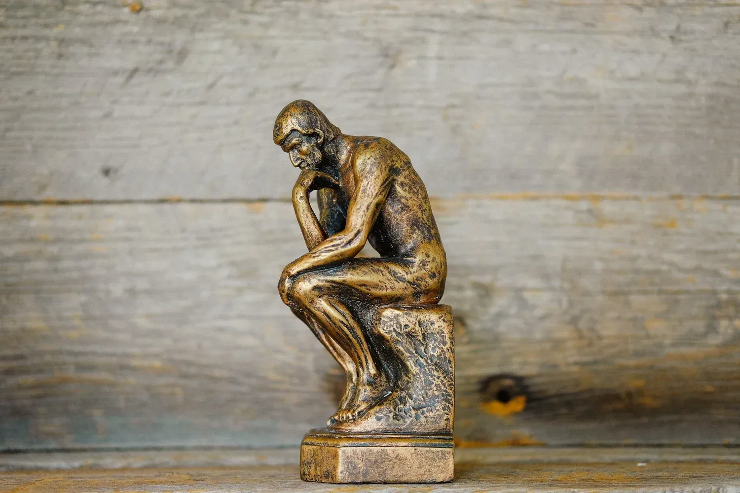 Small bronze Thinker-style sculpture on a wooden surface against a rustic wood background
