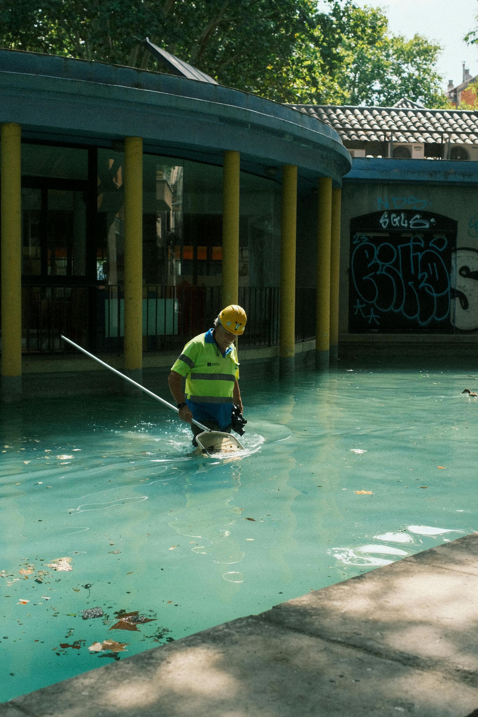Worker in yellow hard hat stands in a shallow turquoise pool beside a curved building with columns.