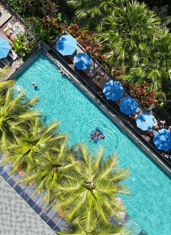 Aerial view of a turquoise resort pool with blue umbrellas, palm trees, and swimmers