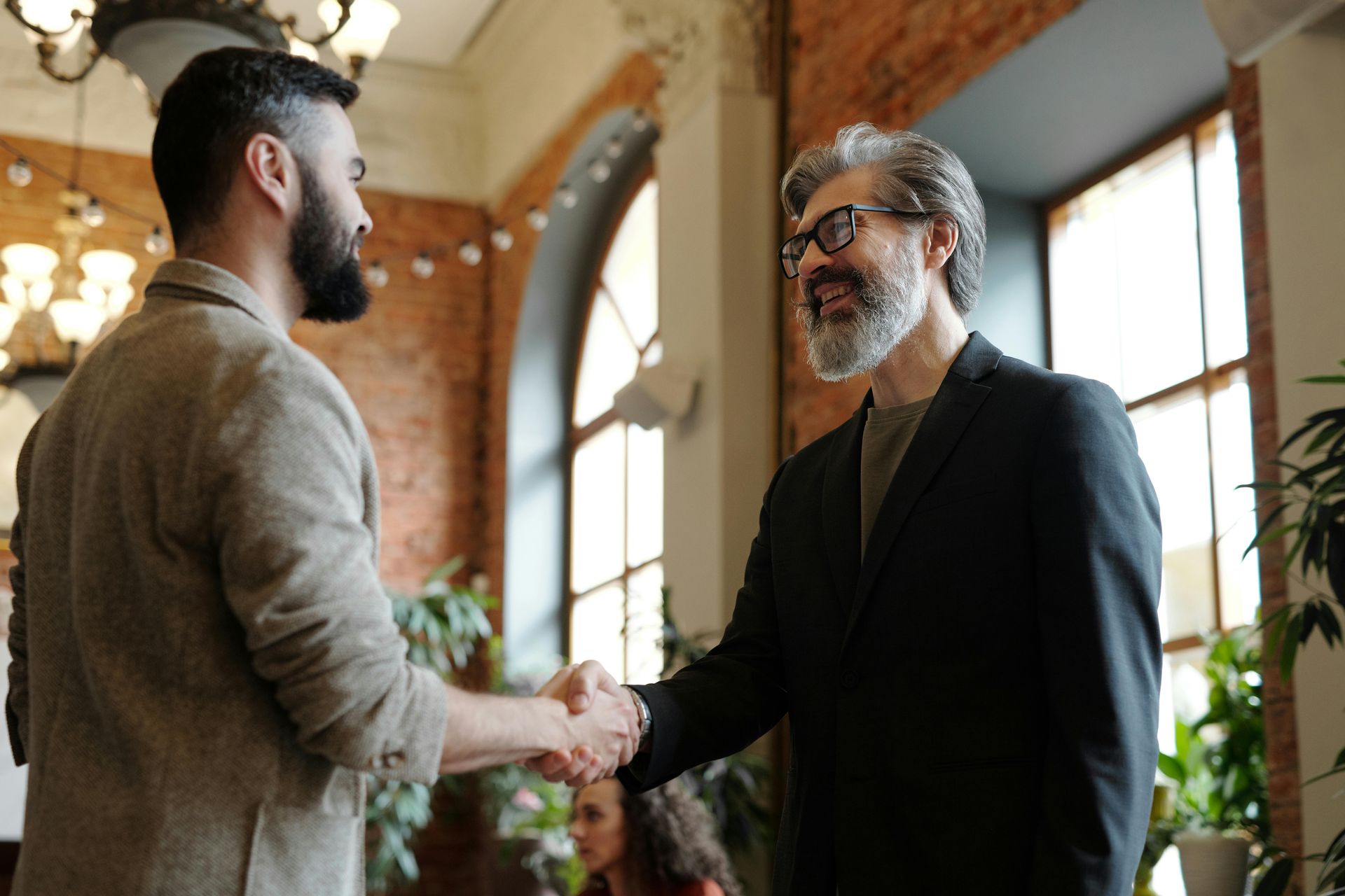 Two men shaking hands in a bright office with large windows and plants.