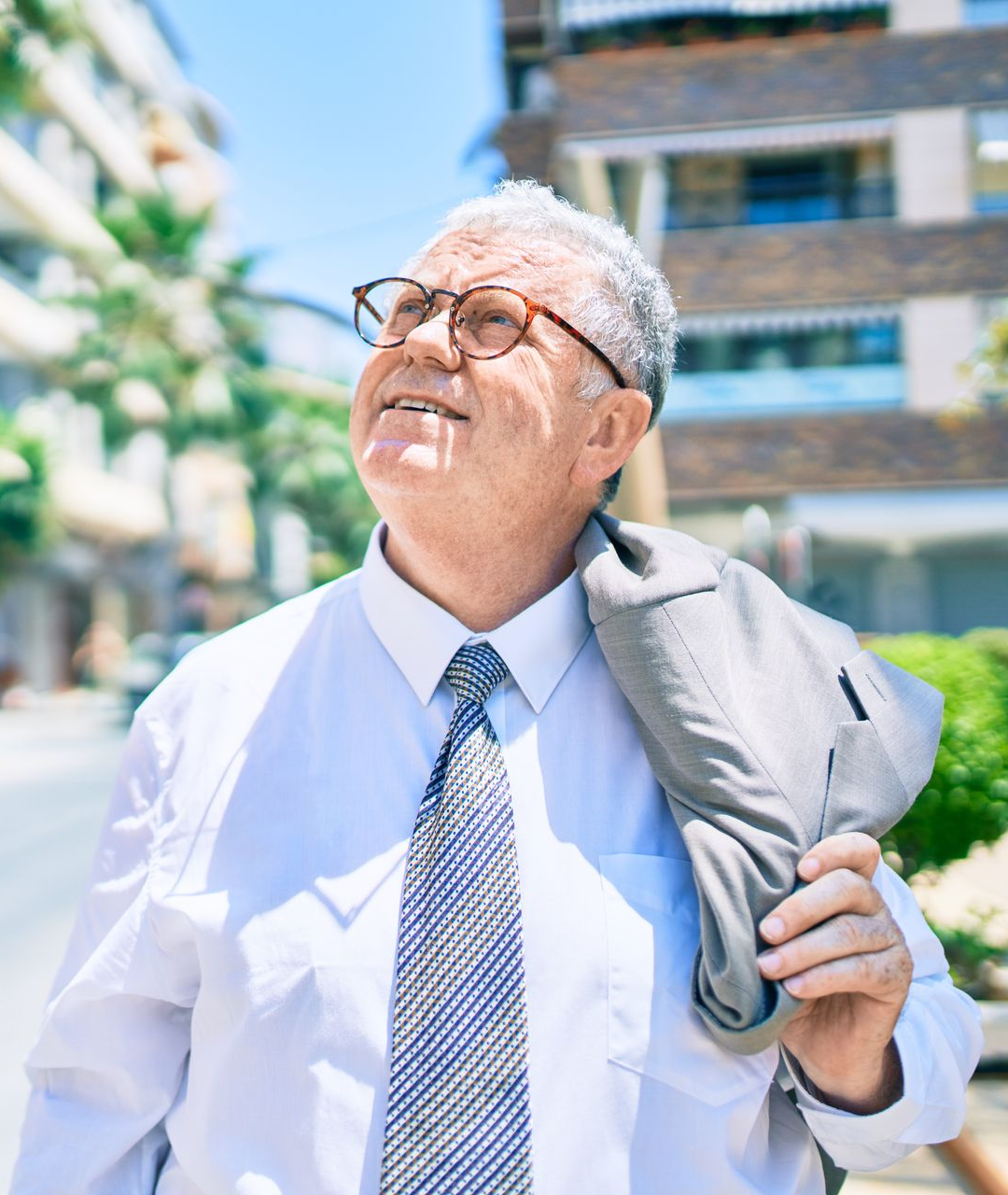 Smiling person in glasses and tie looking up outdoors, holding a jacket over one shoulder.