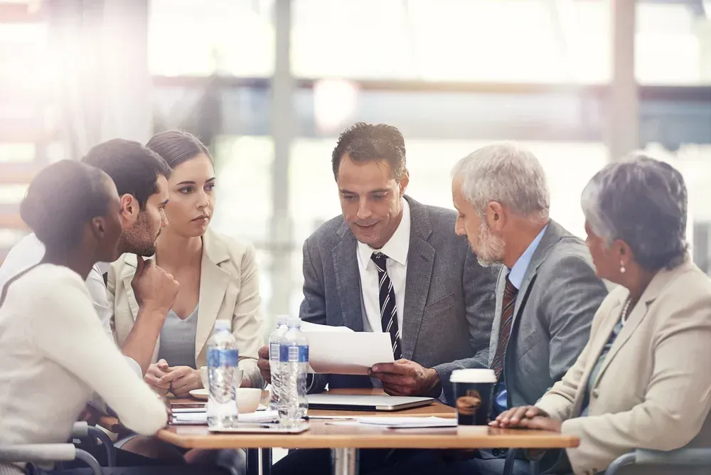 Business meeting with five people seated around a table, discussing documents in a bright office