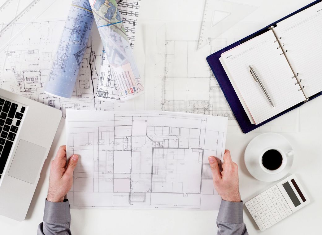 Mani che esaminano planimetrie architettoniche su una scrivania con computer portatile, progetto, quaderno e tazza di caffè.