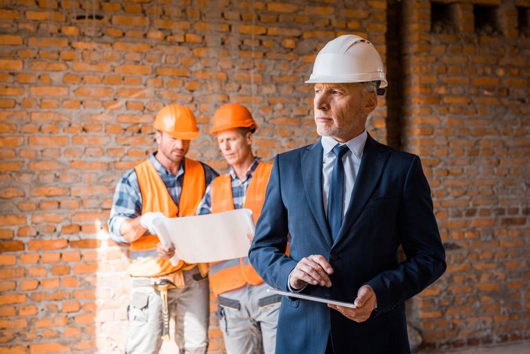 Tre operai edili con elmetti esaminano i progetti in un cantiere con muri di mattoni.