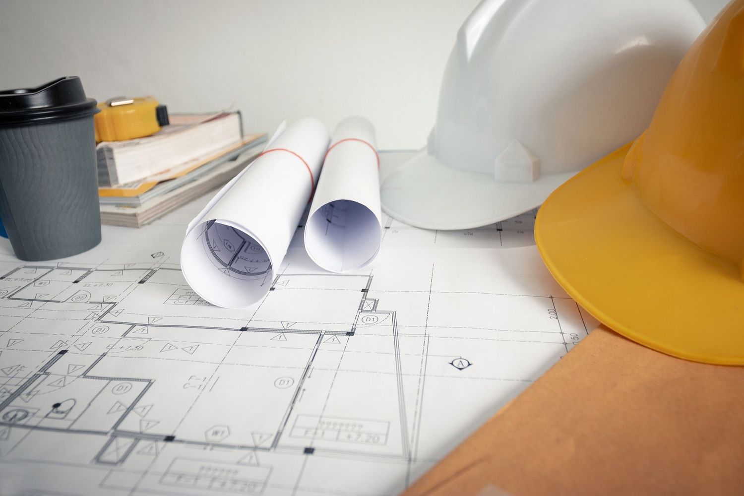 Progetto architettonico con planimetrie arrotolate, caschi di sicurezza e caffè su un tavolo da disegno.