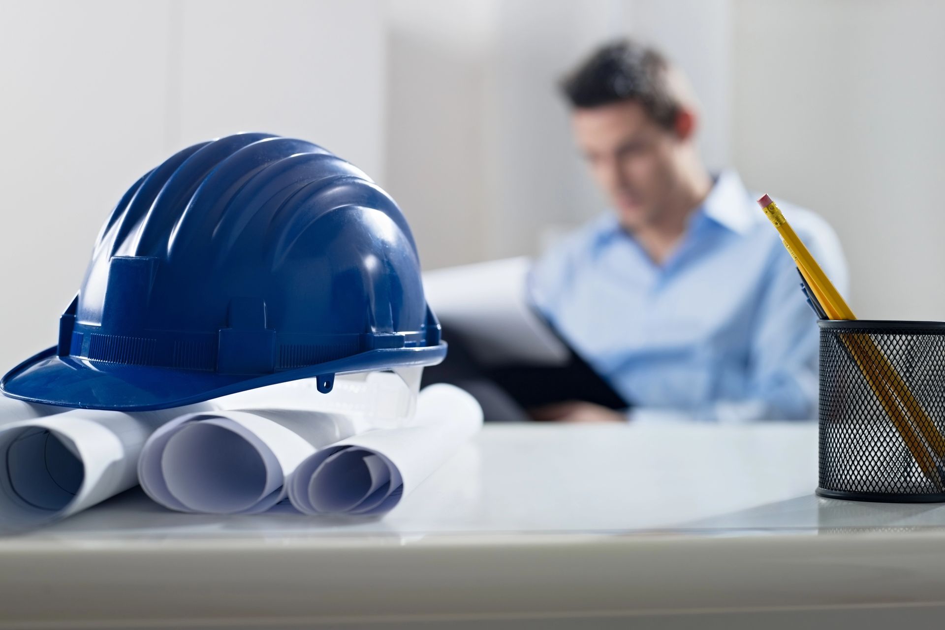 Un elmetto blu e dei progetti arrotolati su una scrivania, con una persona sfocata al lavoro sullo sfondo.