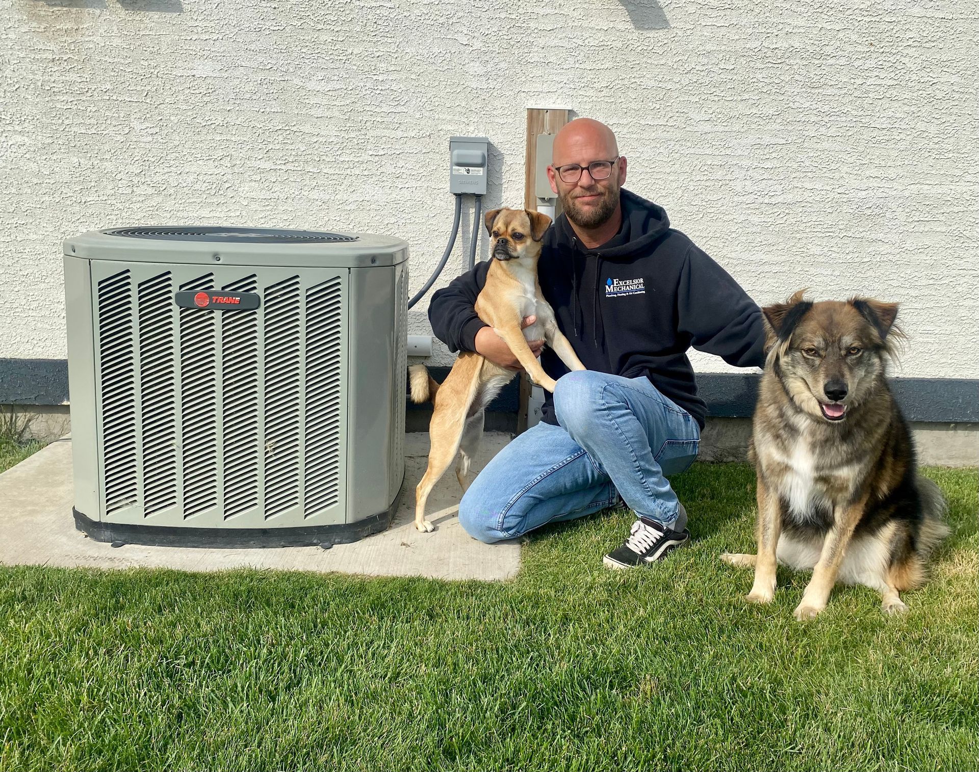Man with two dogs poses by an air conditioner unit on green grass in front of a building.