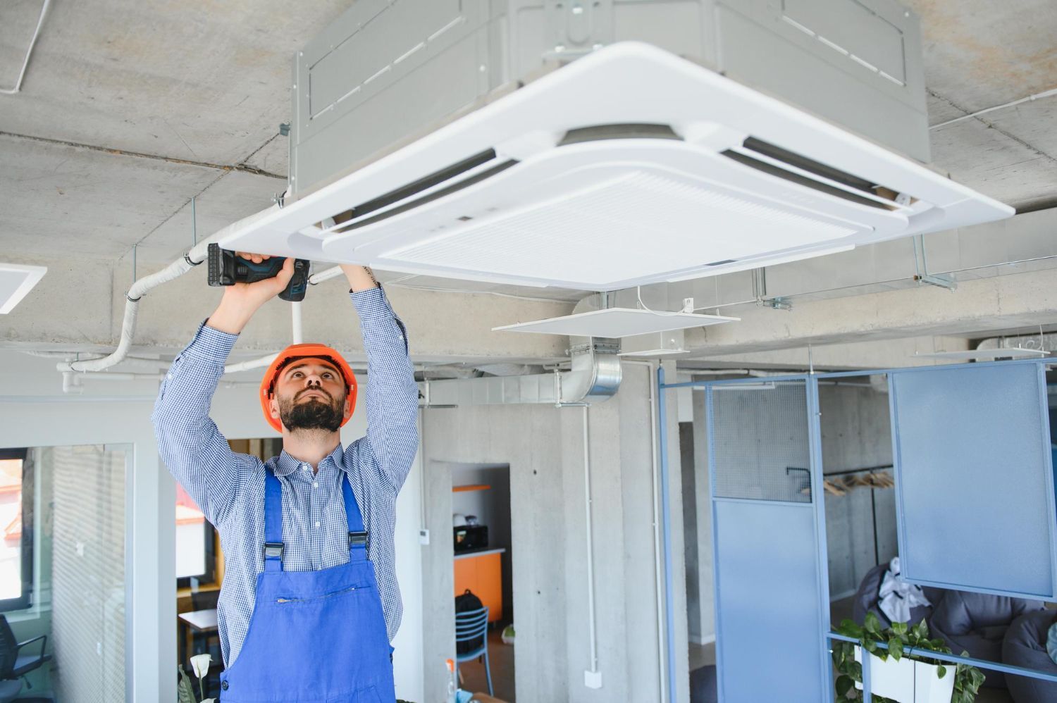 Technician in blue overalls installing a ceiling cassette air conditioner in a bright room