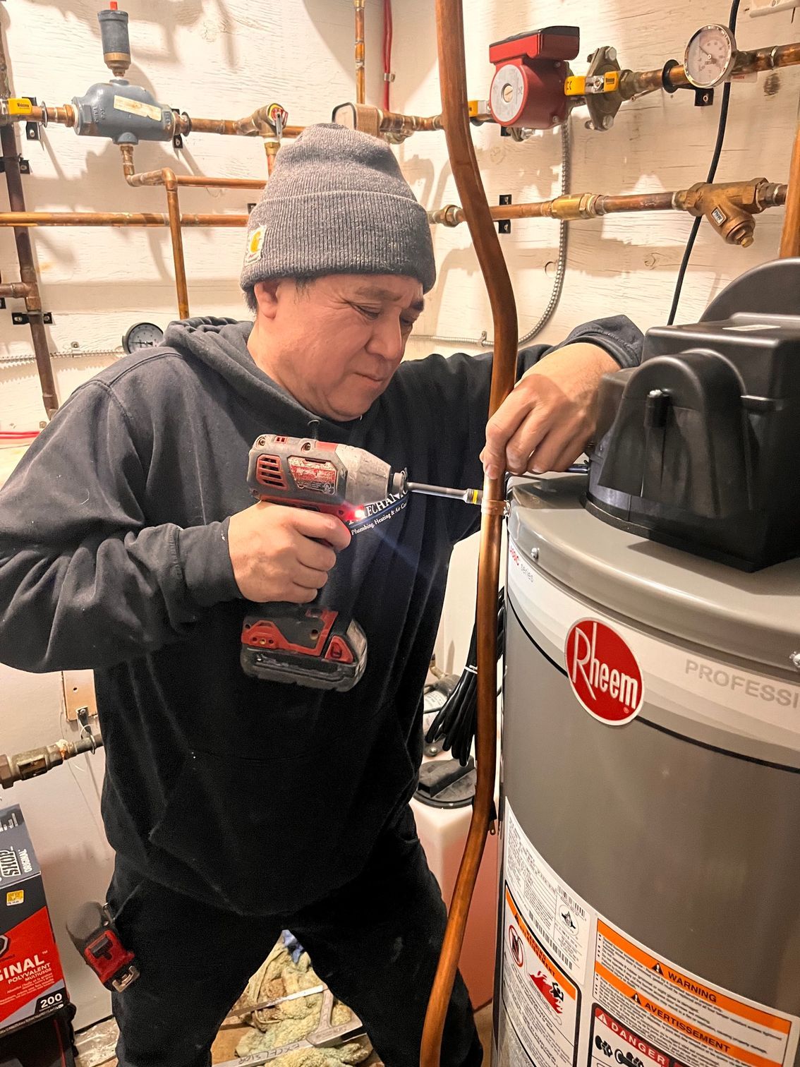 Person in gray beanie using a red drill to install something on a water heater in a workshop.