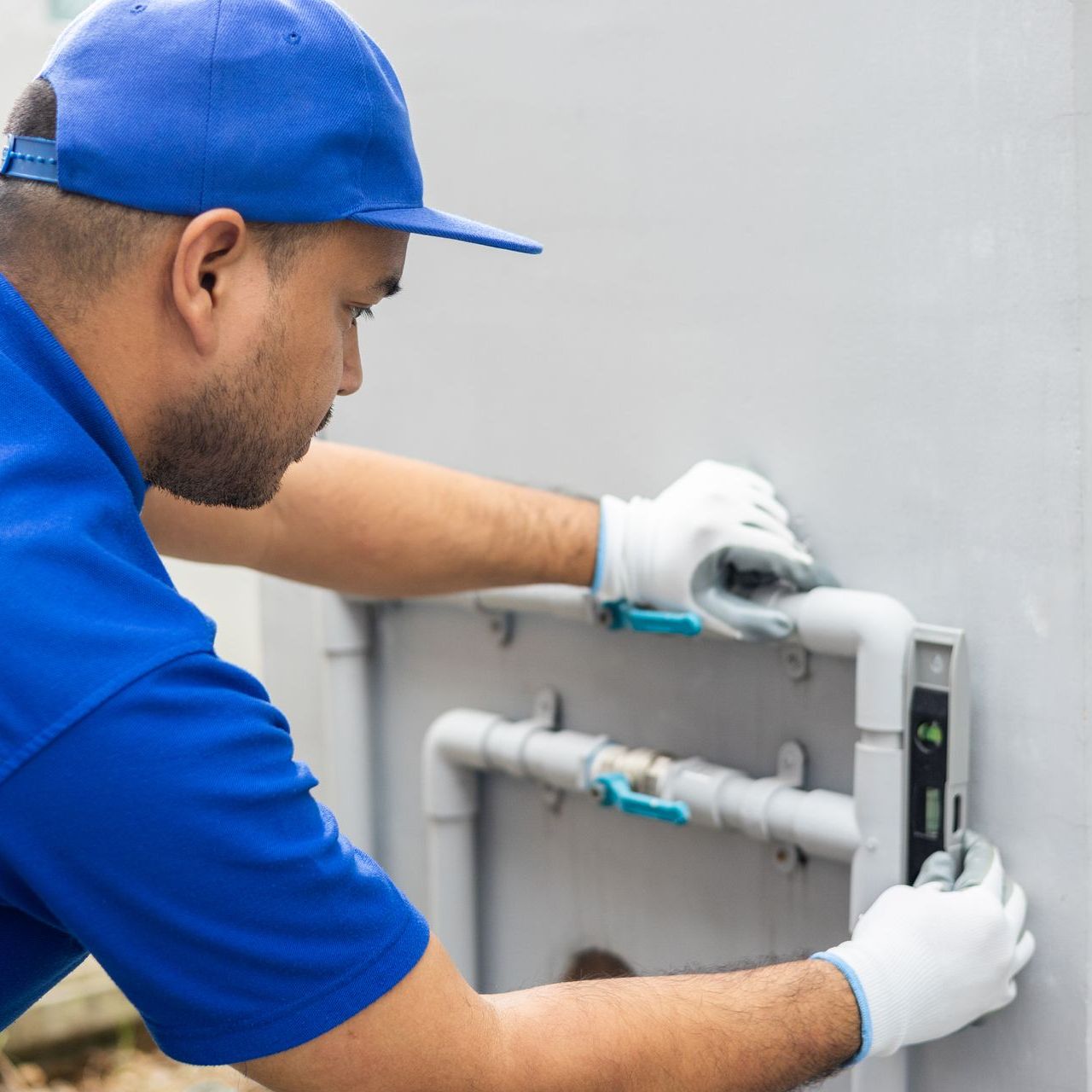 A man in a blue shirt and hat is working on a pipe on a wall.