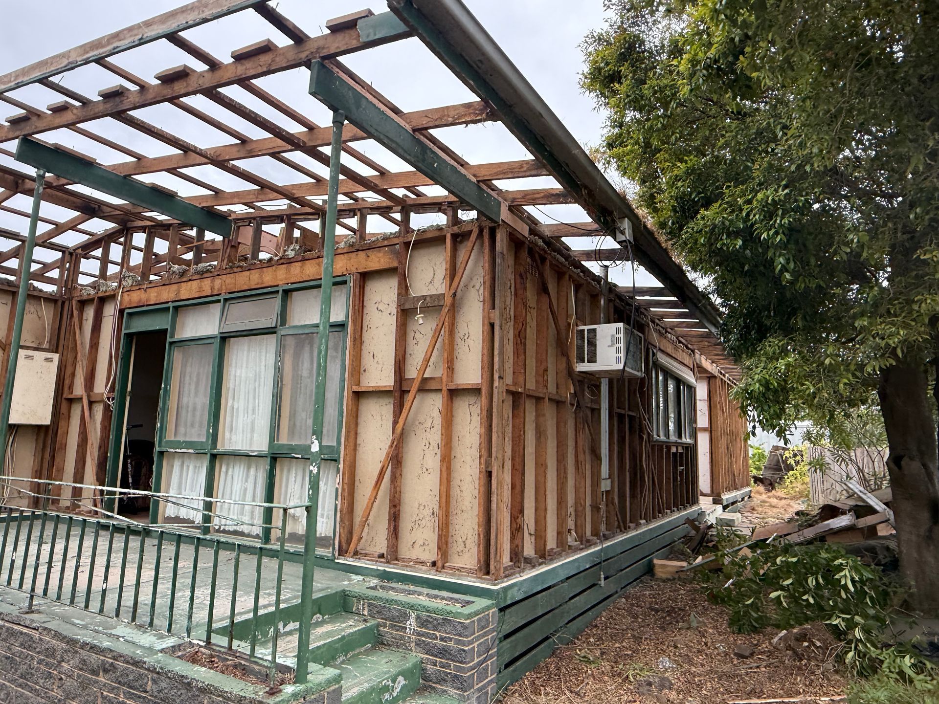 Partially demolished house with exposed wooden frame, green trim, and a tree beside it