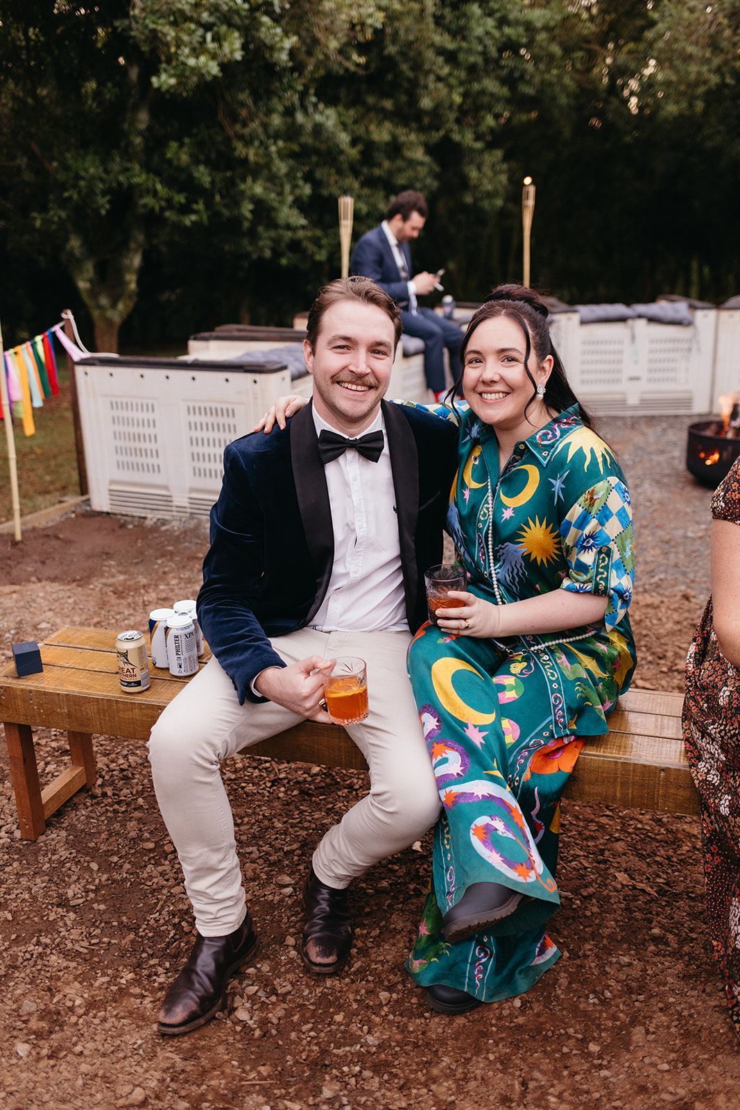 Couple smiling, seated on bench, holding drinks. Man in velvet jacket; woman in patterned outfit. Outdoor setting.