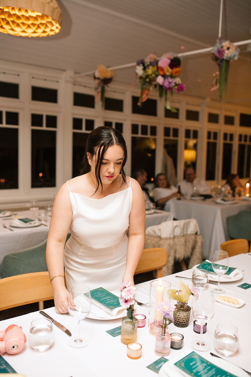 Woman in white dress setting a table at a wedding reception; floral decorations and guests visible.