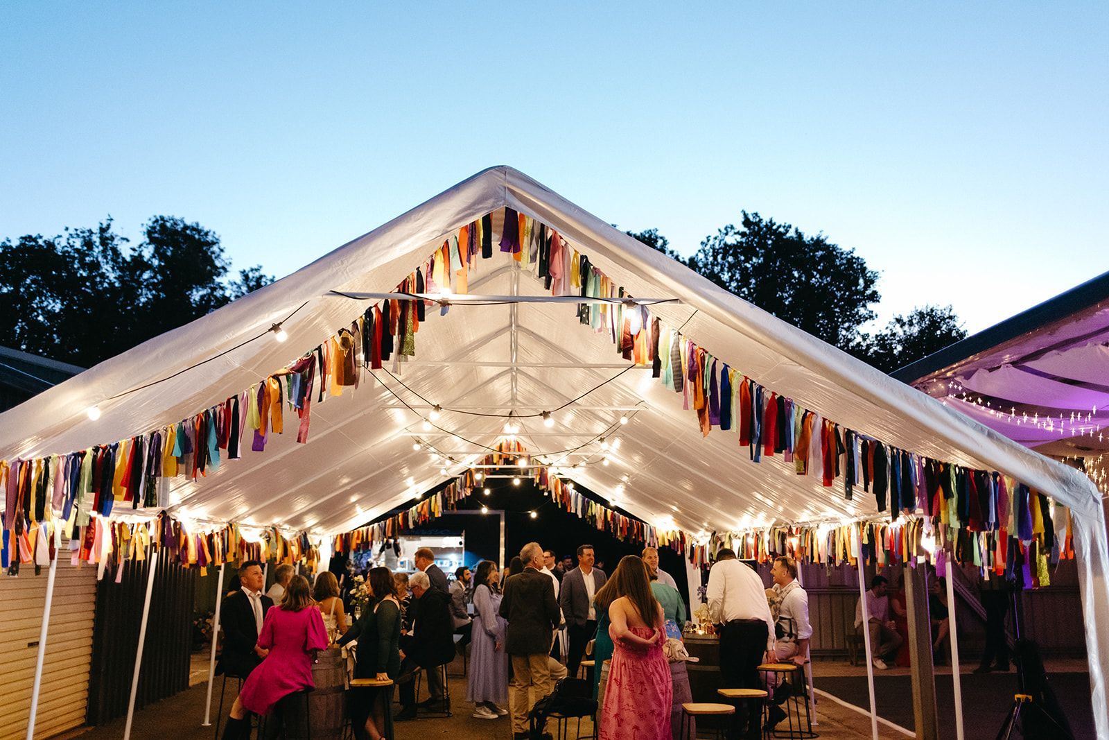 Party tent lit with string lights and colorful fabric strips. Guests mingle, enjoying an outdoor event.