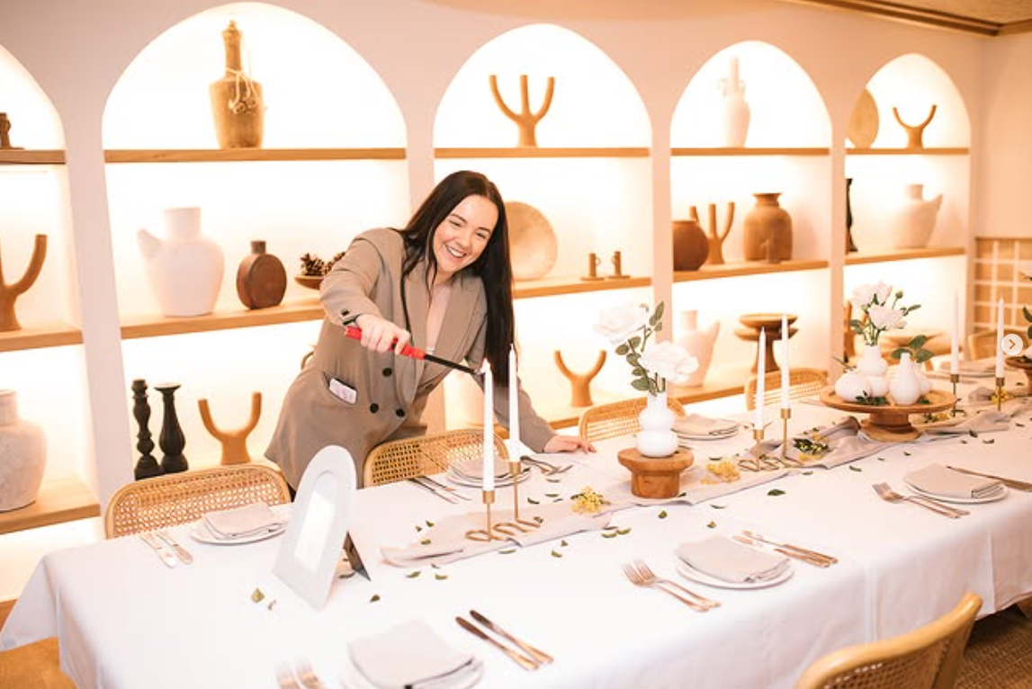 Woman lighting candles on a beautifully set table in a bright room with decorative shelves.
