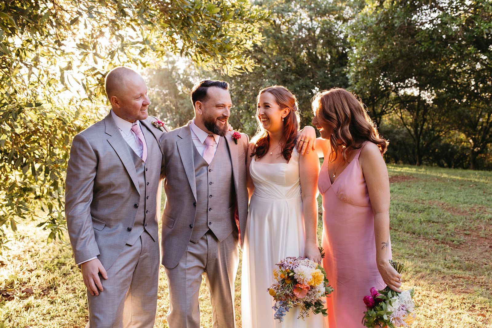 Wedding group, sunny outdoor setting. Groom, bride, and two companions smiling, embracing. Flowers and trees in background.
