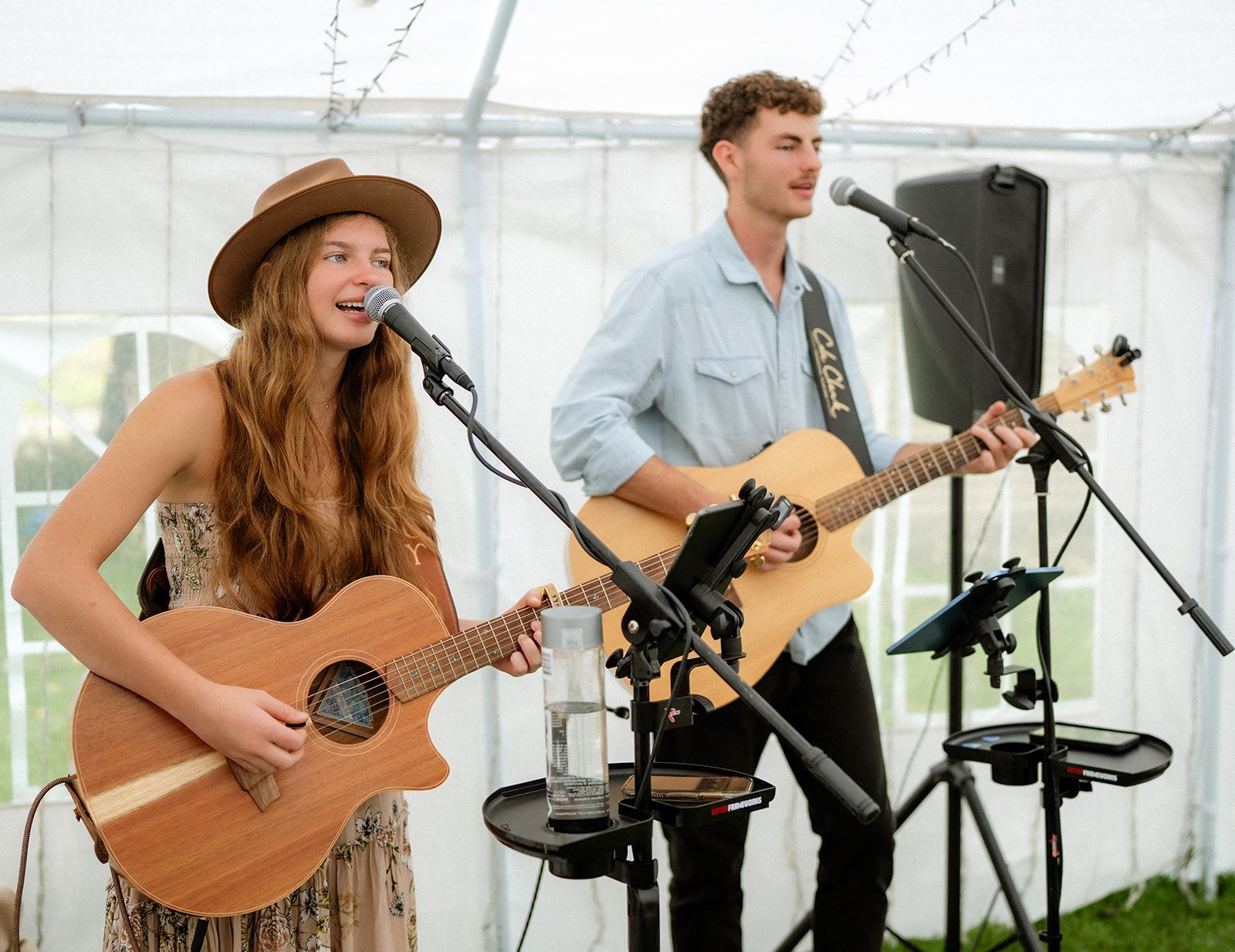 Two musicians performing with acoustic guitars and microphones under a tent.