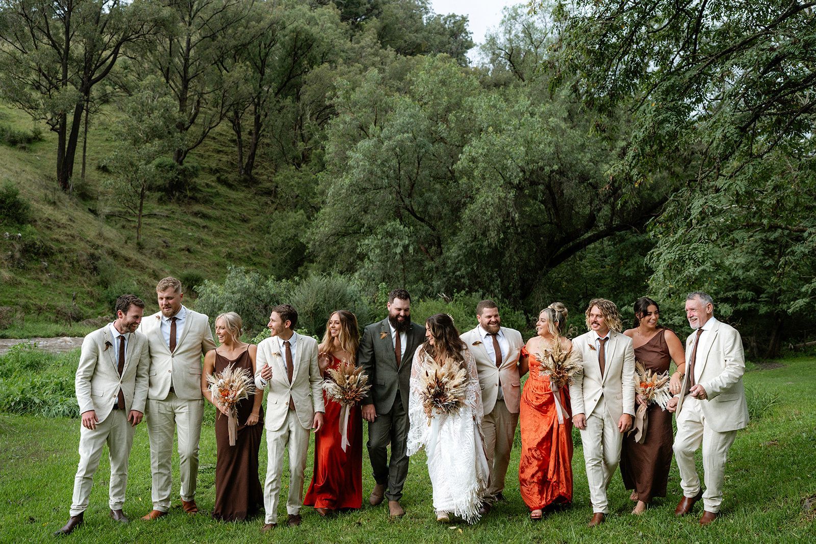 Wedding party in formal attire poses outdoors on grass, green foliage background.