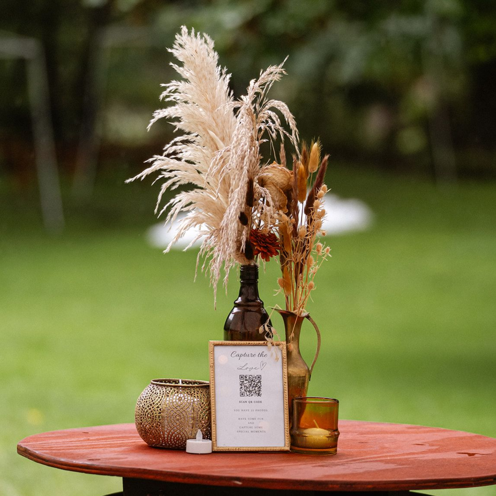Centerpiece with dried flowers in vases on a red table, includes a QR code sign and candles, outdoors.