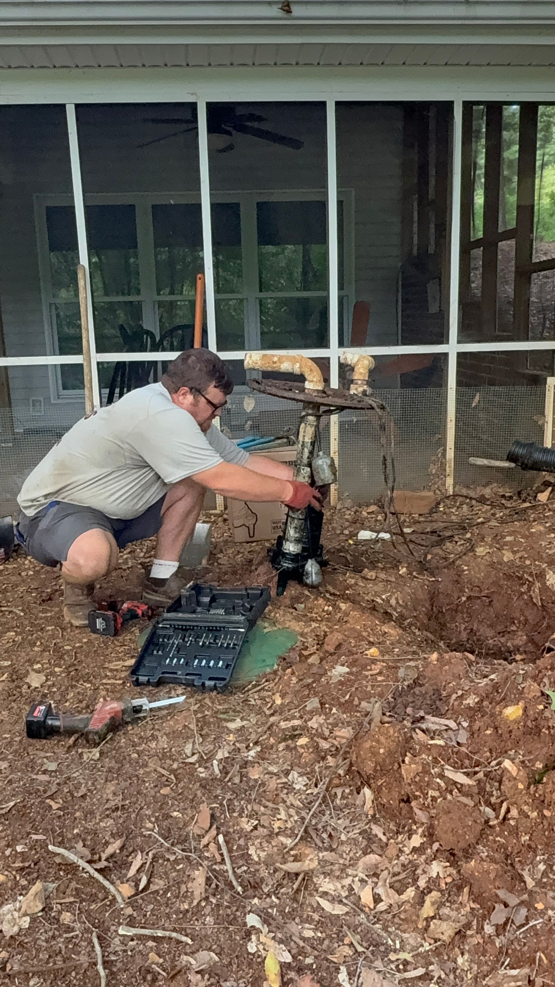Person crouching beside a jackhammer near a house foundation, working in a dirt-filled yard.