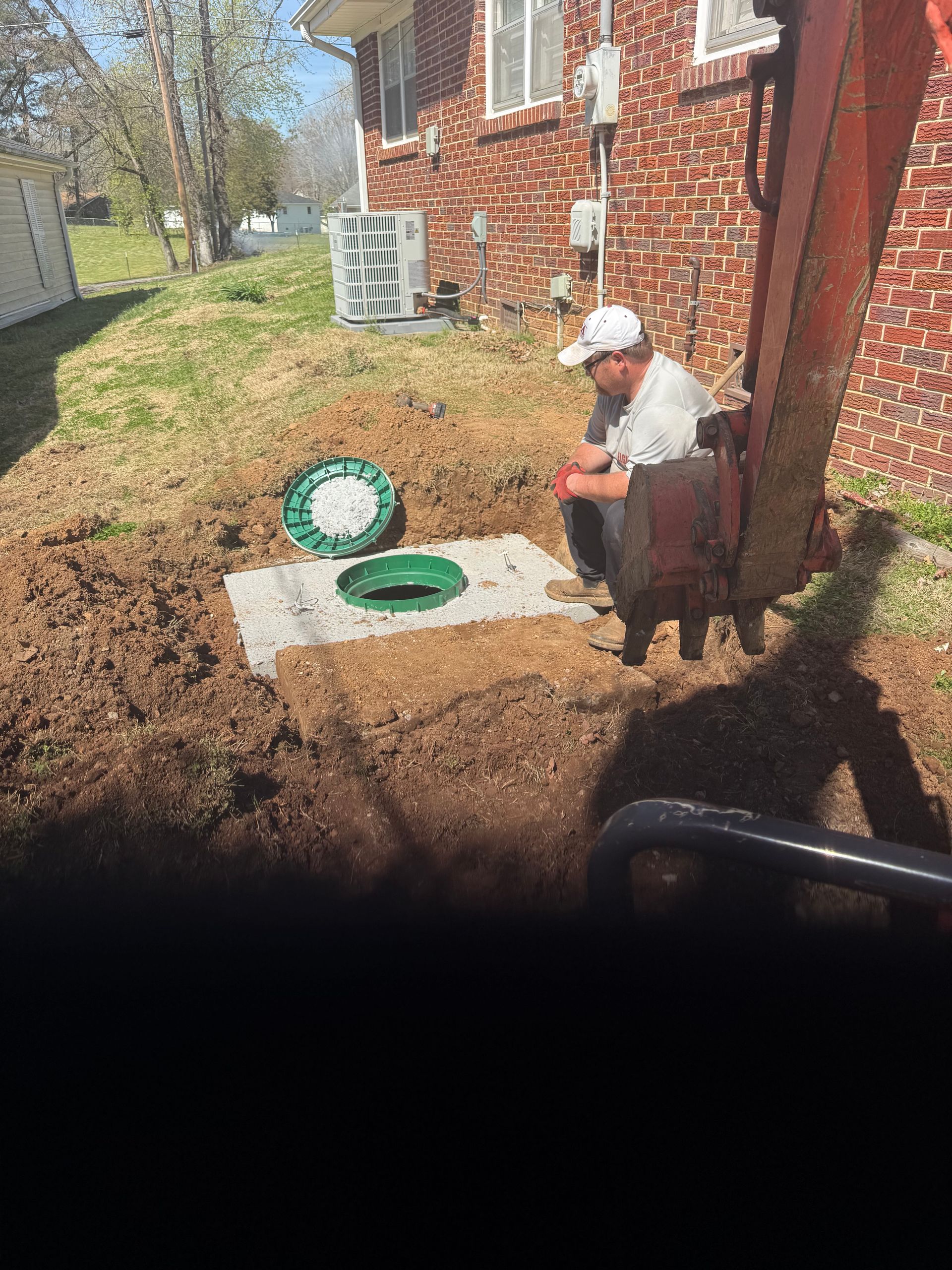 Person sitting by a brick house near two open green utility covers in a yard.