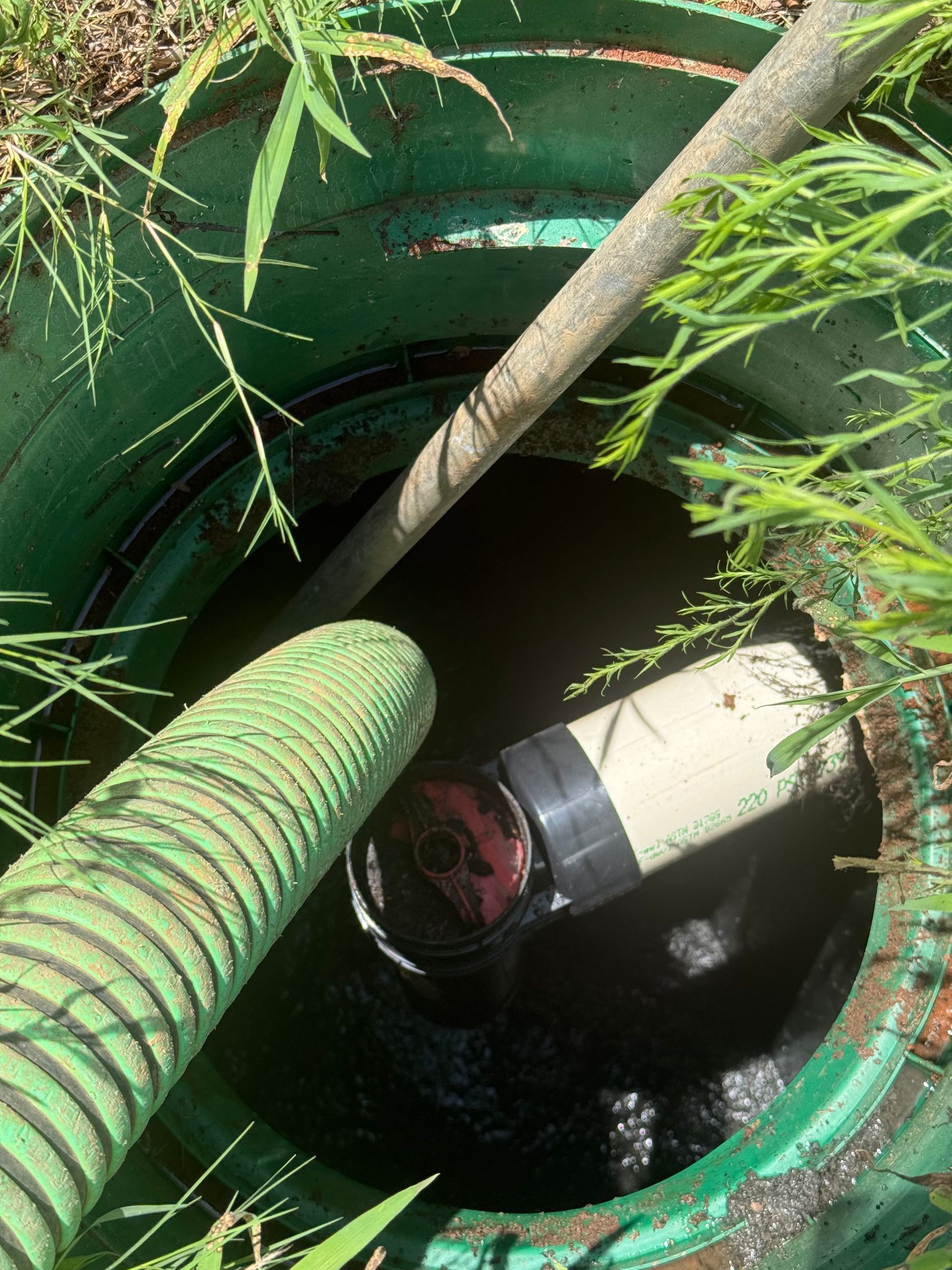 Person in a green utility pit beside a hose and pipe, looking down into a dark opening.