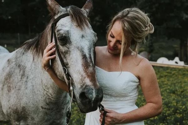 A woman in a wedding dress is petting a horse in a field.