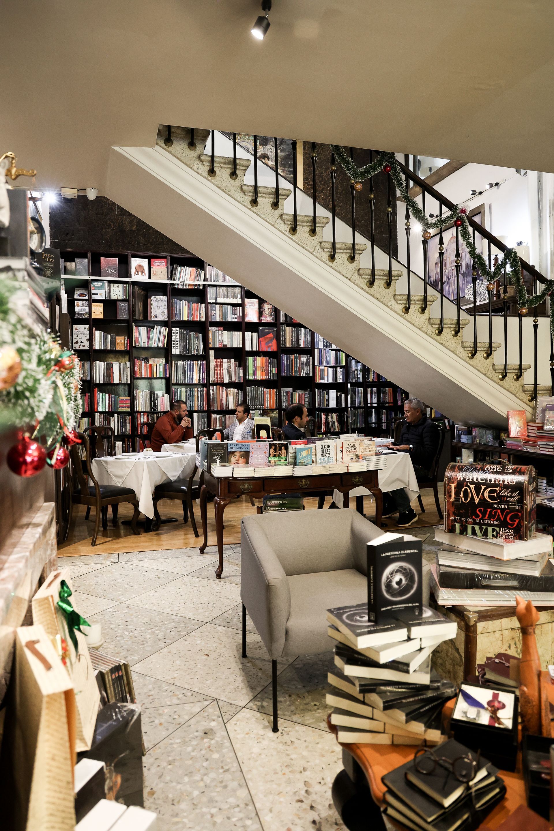 Acogedora librería-cafetería con estanterías, escalera y mesas decoradas, con una iluminación cálida en el interior.